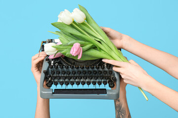 Hands holding vintage typewriter with tulips on blue background. Women's Day celebration