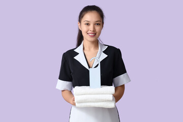 Young Asian chambermaid with clean towels and door hanger on lilac background