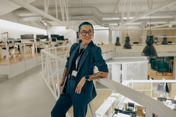 Smiling tattooed female manager with green short hair holding laptop standing in modern office