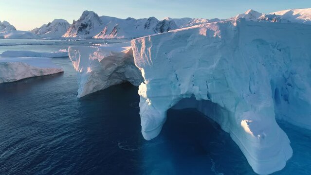 Huge icebergs blue cave, melting glacier in Antarctica. Large mass of ice floating polar ocean water. Snow covered mountains in pink sunset light. Antarctica travel, Arctic landscape. Drone flight