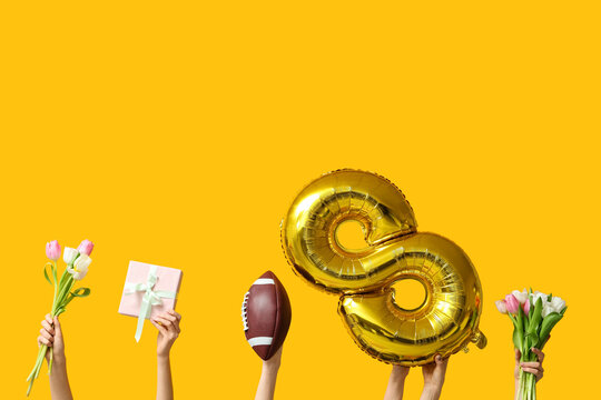 Female hands with foil balloon in shape of figure 8, rugby ball and tulip flowers on yellow background. International Women's Day - Powered by Adobe