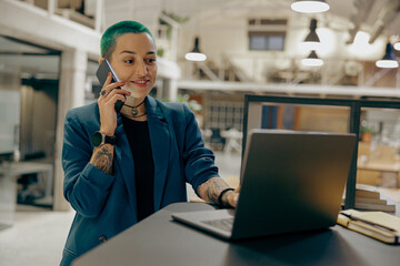 Stylish tattooed female business owner is use laptop while talking phone sitting in coworking