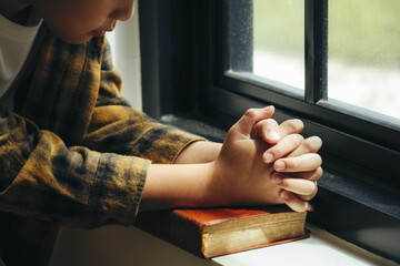 A boy folded hands on holy bible by the window in library and church, religion and worship concept.