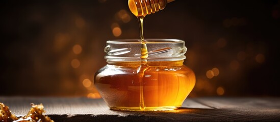 A glass jar filled with golden honey stands on a wooden table, next to a honey dipper. The honey is slowly dripping from the dipper into the jar, creating a warm and inviting scene. - obrazy, fototapety, plakaty