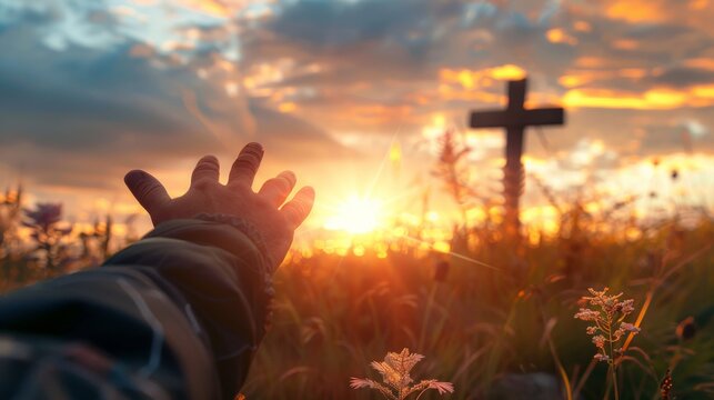 A person's hand reaching out towards the sun during a vibrant sunset with a cross in the background.