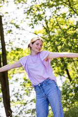 The image portrays a youthful woman mid-dance, her arms outstretched as if to welcome the embrace of a summer breeze. She wears a lilac-colored top, complementing the lively greens of the trees in the