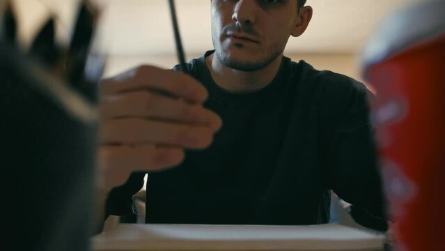 The camera frames an artist between a coffee cup and pencil holder as he selects his pencil and the introspective moment of creative deliberation, as he gazes down at his sketchpad.