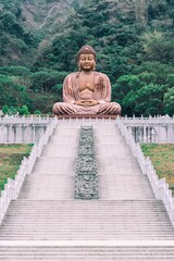 Visualize a colossal statue of the Buddha seated serenely at the base of a towering mountain, its serene countenance radiating a sense of peace and tranquility that envelops the surrounding landscape. - obrazy, fototapety, plakaty