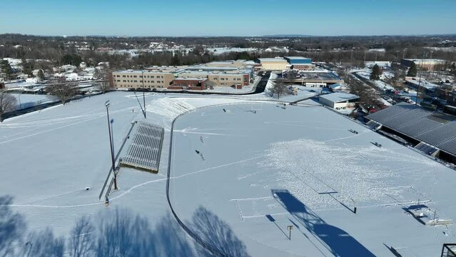 Aerial shot of a high school sports field covered in snow. Football stadium and track with bleacher seating. Winter in America theme.
