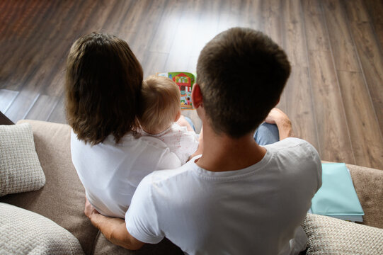 Parents read a book to their child, sitting on the sofa, looking at educational pictures, rear view. Young loving parents spend time with their baby. Concept of happy family, learning
