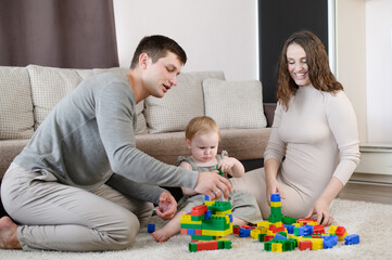 Obraz na płótnie Canvas Young family: dad, mom playing with a child with a multicolor plastic construction set,sitting on floor at home,having fun together on weekend, family vacation. Educational game for children, leisure