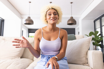 Young biracial woman with blond curly hair sits on a beige sofa, on a video call