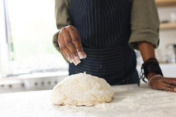 A young African American woman sprinkles flour over dough on a kitchen counter with copy space