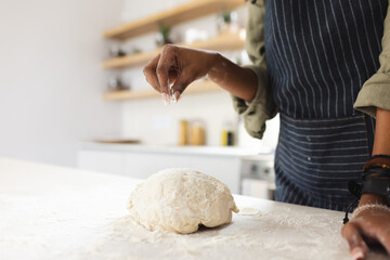 A young African American woman sprinkles flour over a ball of dough on a kitchen counter with copy s