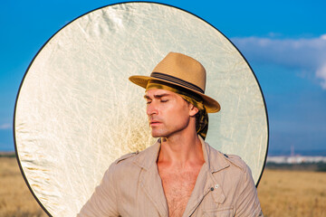 Portrait of a tall handsome man dressed in a coarse linen suit and hat standing at golden oat field with a golden reflector on the background.