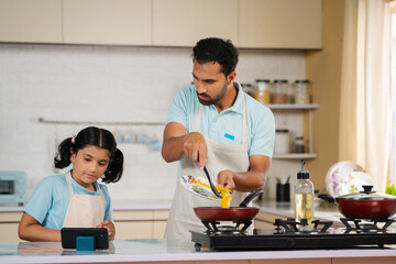 curious father with daughter cooking together by watching online video from mobile phone at kitchen - concept of family bonding, weekend activities and teamwork