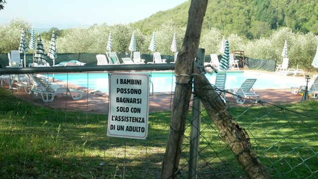 Pan left of a public pool with a fence sign that reads "Children Can Only Bathe With The Assistance Of An Adult" in Italian