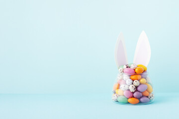 Colorful candy jar decorated with bunny ears against blue background, gifts for Easter