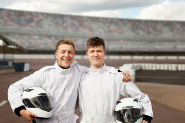 Two friends ready for a race, smiling with helmets in hand at a racetrack. 