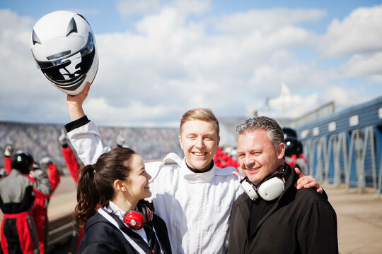 A joyful trio celebrates at a racetrack, with one person holding a helmet aloft triumphantly. 