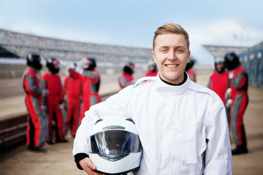 Smiling racer holding helmet with team in background at the track.  