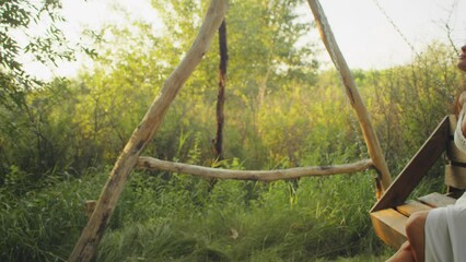 Woman in a white dress relaxing on a wooden swing in a lush forest at golden hour. Peaceful solitude and nature enjoyment concept. Design for wellness retreats, lifestyle blogs, and eco-friendly