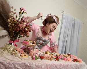 Close up portrait of cute female model wearing an opulent pink gown, the costume of a historical French baroque nobility. Eating indulgent food feast with pastries, flowers and rich jewels