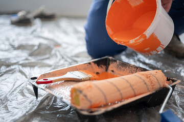 Room renovations at home concept. Close-Up of man pouring orange paint on a roller and tray. Paint...