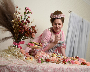 Close up portrait of cute female model wearing an opulent pink gown, the costume of a historical French baroque nobility. Eating indulgent food feast with pastries, flowers and rich jewels