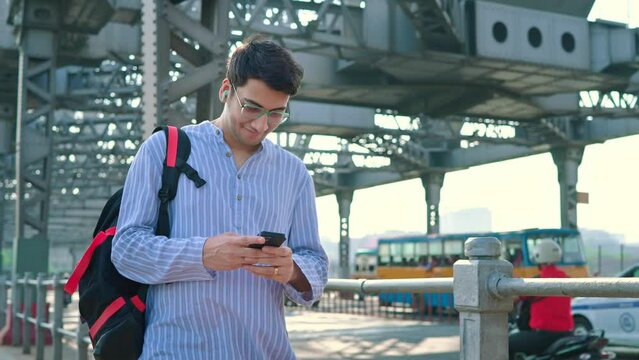 An attractive smiling Indian Asian male or man is carrying a backpack on his shoulder using a mobile phone and walking on an iconic Howrah bridge in Kolkata with vehicles passing in the background