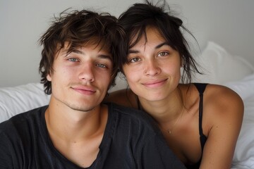 A man and a woman sit cheek to cheek on a bed, smiling with matching hairstyles