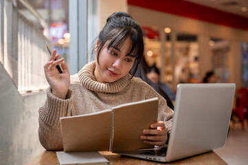 A beautiful and positive Asian woman in a cosy sweater is working remotely at a coffee shop.