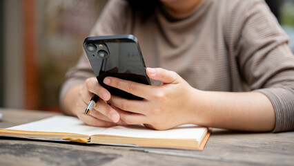 A cropped shot of an Asian woman using her smartphone while reading a book or doing some work.