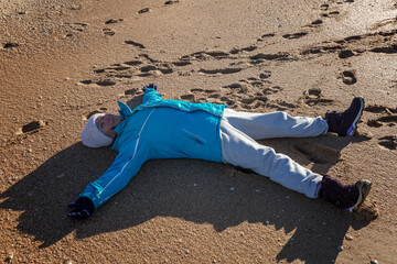Smiling elderly woman lying on the sand in a star pose wearing a jacket and hat during the winter season. Active life in adulthood.