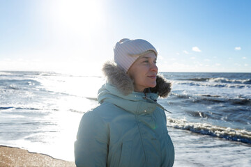 A young woman in a blue jacket walks along the seashore in the winter season on a sunny day. Active lifestyle and walks in nature. Close-up.