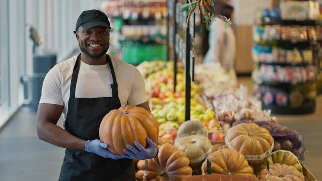 Portrait of a happy man with Black skin color in a white T-shirt near a display case with pumpkins a man holds a pumpkin in his hands poses and smiles broadly in a large grocery store