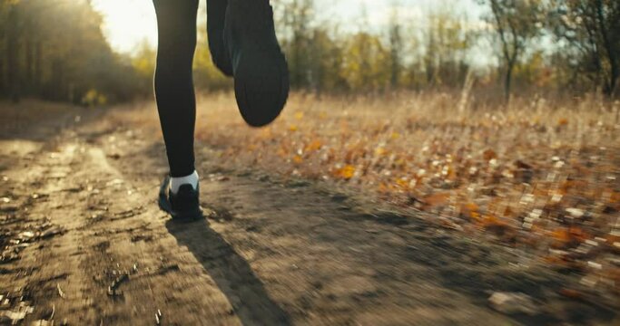 Close up a man in a black sports uniform and black sneakers runs along an earthen path in the autumn forest among fallen brown leaves and dry grass in the fall