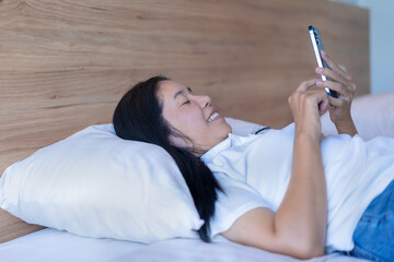 Beautiful Asian woman enjoying playing mobile phone over wireless network in bedroom. Talk with your lover or close friend, and shop online. Look happy, show a smile, and surf the internet.