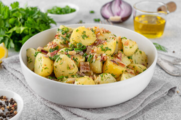 Warm potato salad with bacon, onion, fresh herbs and vinegar dressing in a bowl on a gray concrete...