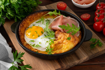 Savory Dutch baby pancake with cheese, fried egg, arugula and prosciutto in cast iron pan on a dark wooden background.