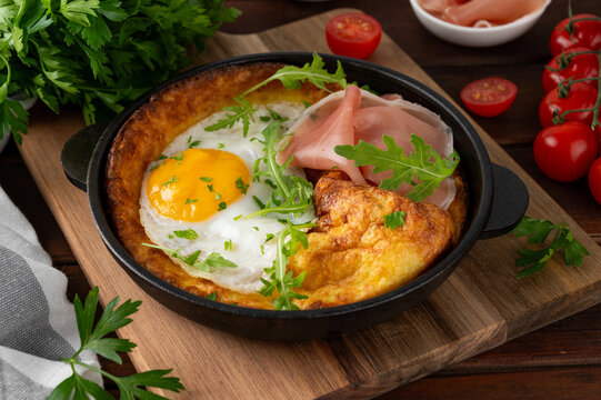 Savory Dutch baby pancake with cheese, fried egg, arugula and prosciutto in cast iron pan on a dark wooden background.