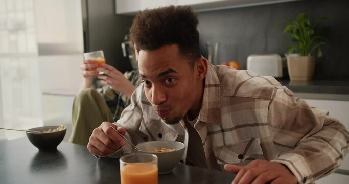 A young man with Black skin color in a cream plaid shirt does not like the cereal with juice that he eats from his bowl and he spits it right there during his breakfast in a modern apartment with his