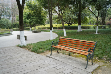 A park bench, standing silently in an environment surrounded by lush trees. The area around the bench is covered in grass and additional trees, 