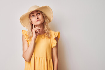 Woman in yellow dress and straw hat looking thoughtful against a light background.