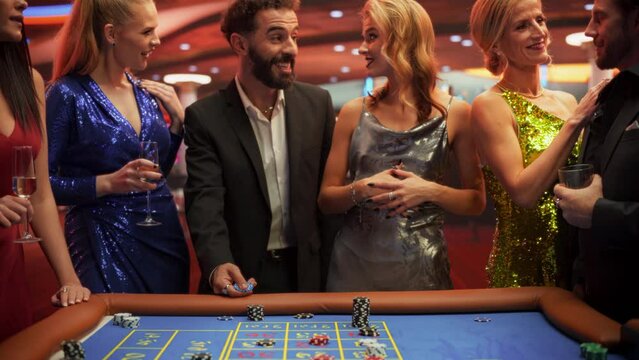 Cheerful Group of Rich Young People Gathered Around a Roulette Table at a Modern Casino. Cinematic Footage of a Handsome Hispanic Male Throwing a 5000 Dollar Casino Coin Towards the Camera
