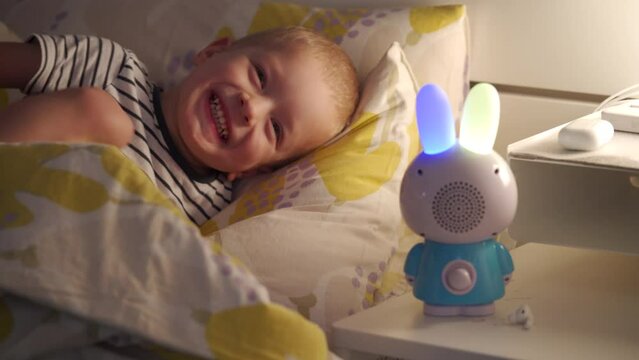 happy child playing with interactive musical toy smart bunny lying in bed in the evening. Boy listening to bedtime story.