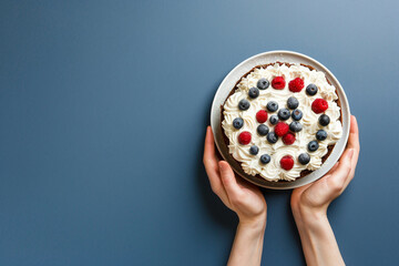 Woman hands holding sponge cake decorated whipped cream, raspberries and blueberries on a blue background. Copy space. Top view. - obrazy, fototapety, plakaty