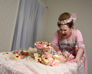 Close up portrait of cute female model wearing an opulent pink gown, the costume of a historical French baroque nobility.  sitting at table with sweet cakes and indulgent food feast.