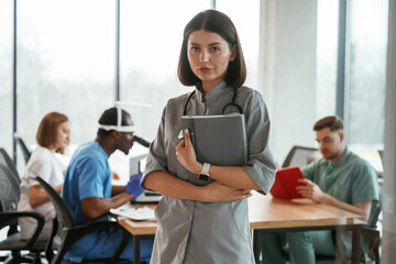 Woman in grey uniform is standing with notepad. Group of doctors are together indoors