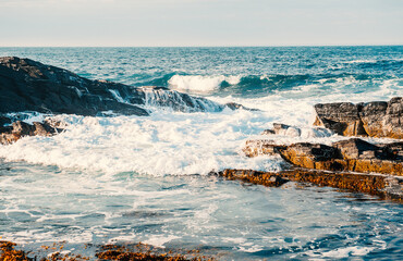 Sea waves crashing on rocks in sunny day against cloudy sky
